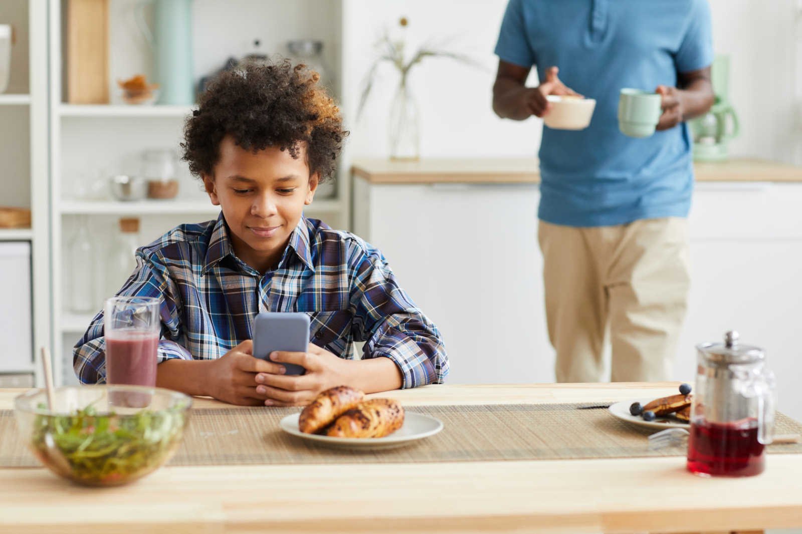 A boy at a kitchen table looks at a smartphone while an adult stands behind him holding a bowl and a mug.
