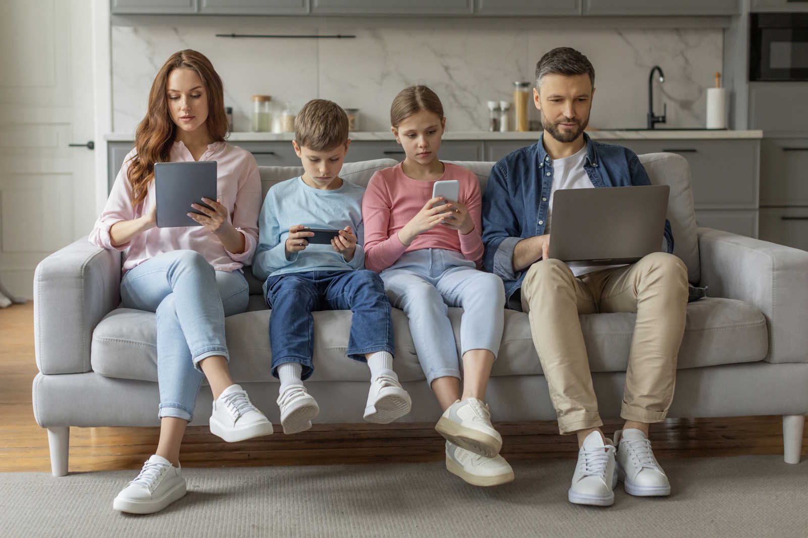 Two adults and two children sit in a row on a sofa, each looking at a different device: a tablet, a handheld games device, a phone and a laptop.