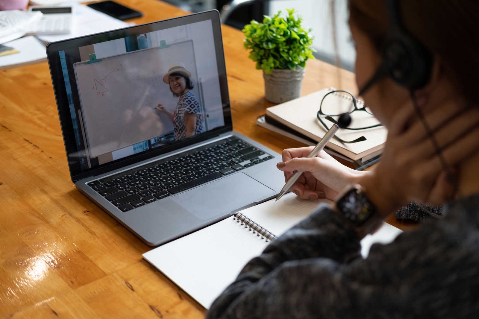 An adult learner wearing a headset takes handwritten notes beside a laptop, with a teacher visible on the screen standing at a whiteboard.