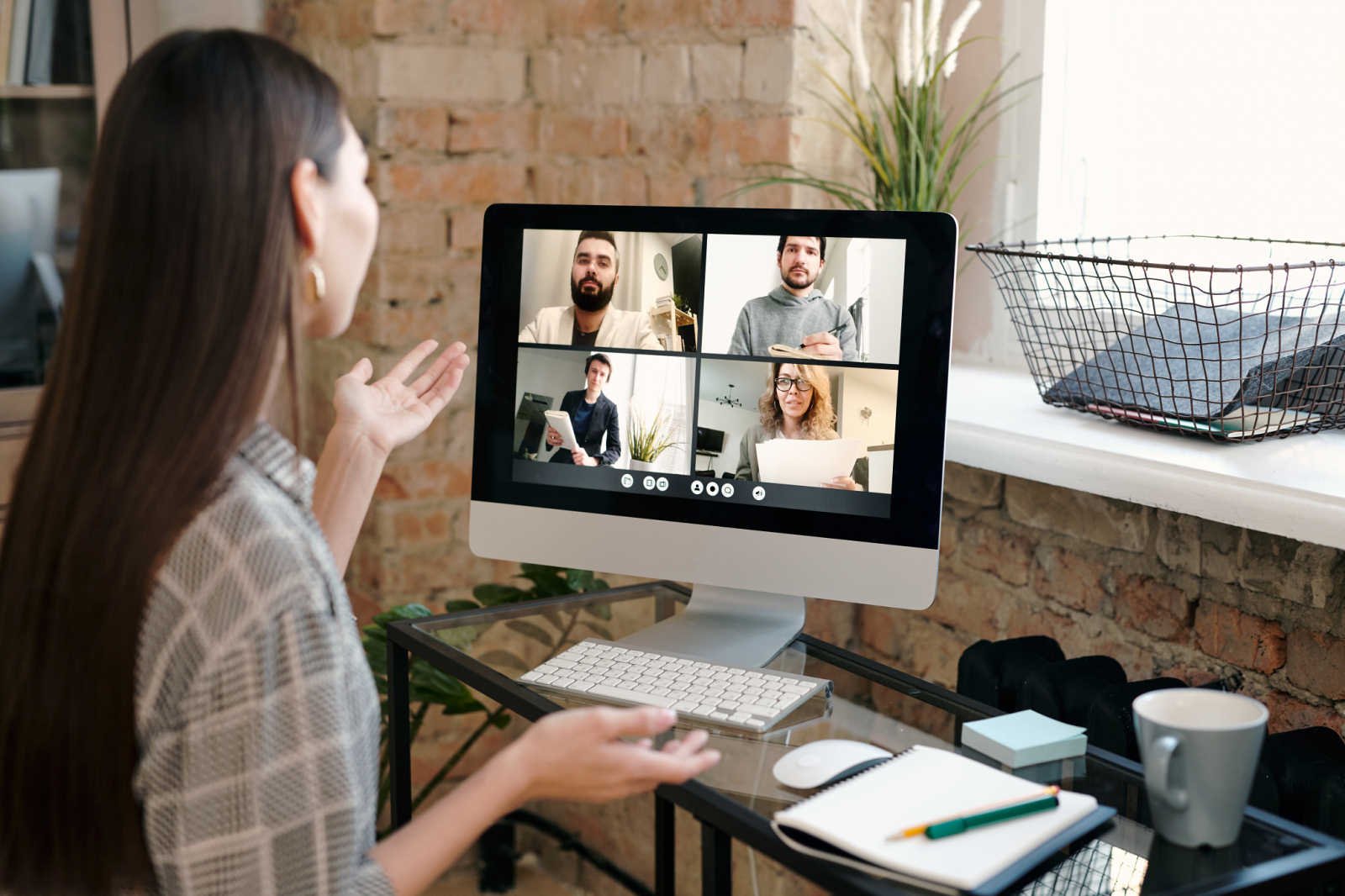 A woman speaks and gestures at a desktop monitor showing four other participants in a video call grid.
